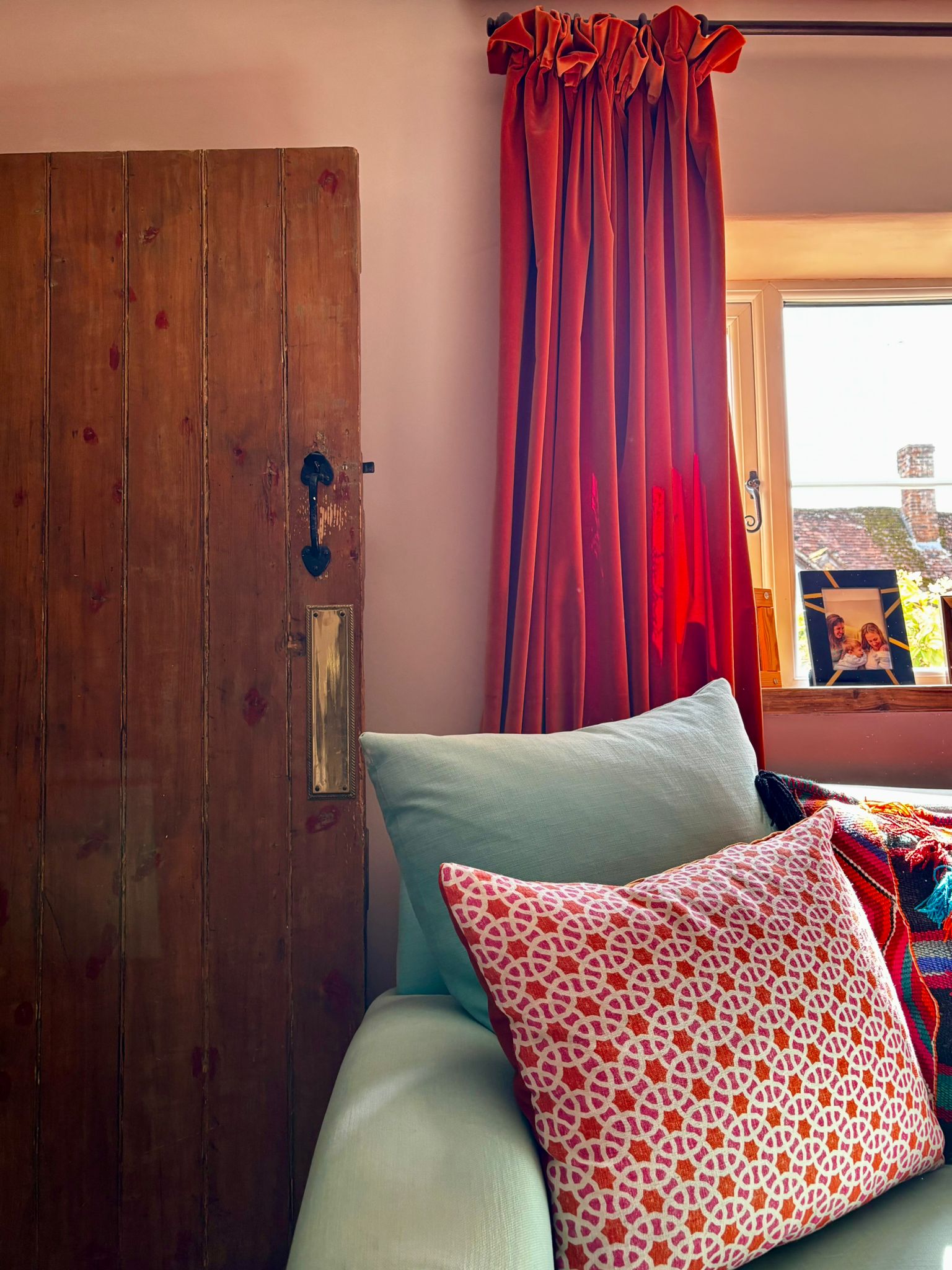 Layered living room interior with rustic wooden door, deep red velvet curtains and sage green sofa with patterned cushion
