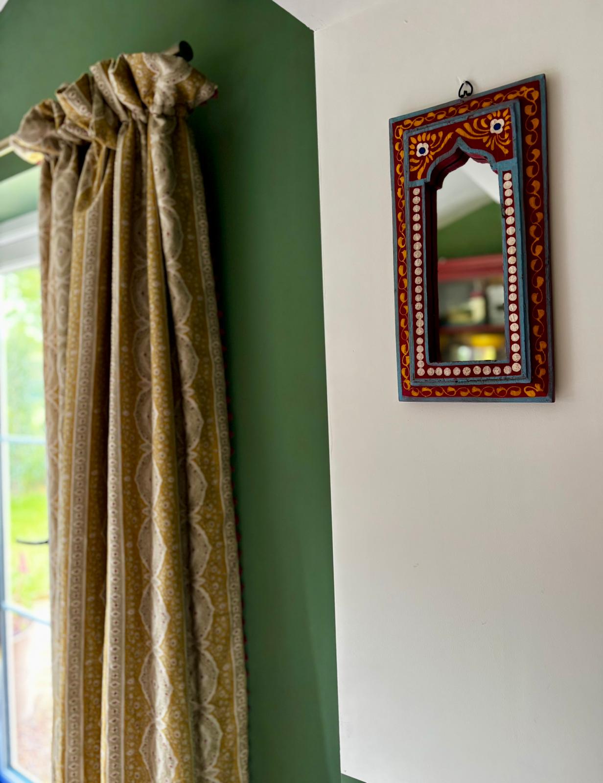 Hand-painted Rajasthani-style mirror on a deep green wall beside a soft ochre block-print curtain